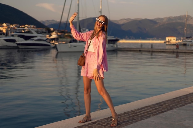 日没時にモンテネグロマリーナでポーズをとってトレンディなピンクのリネンのスーツを着ている女性のアウトドアファッションの肖像画は彼女の贅沢な休暇をお楽しみください