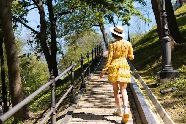 Открытый модный портрет женщины в желтом летнем платье и шляпе, идущей по тропинке в парке, вид сзади