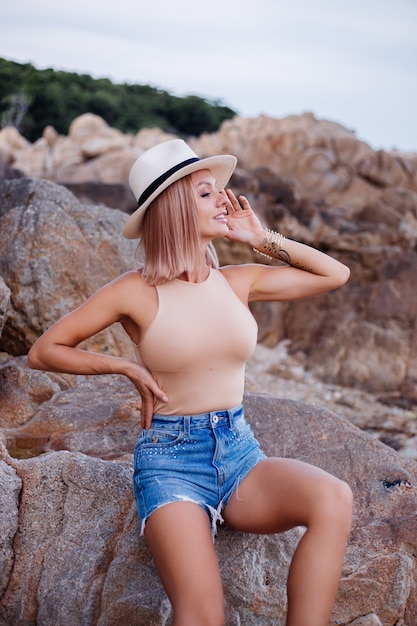 Ritratto di moda all'aperto di giovane donna europea in pantaloncini di jeans blu body corpo beige e cappello elegante classico sulla luce del tramonto calda spiaggia rocciosa tropicale