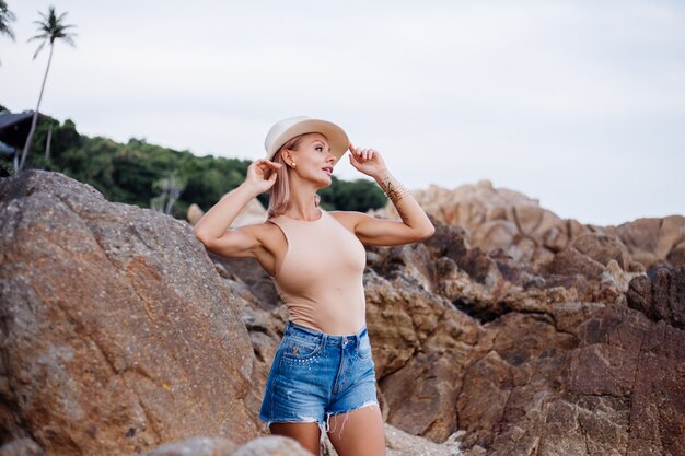 Outdoor fashion portrait of young european woman in beige body bodysuit blue jean shorts and classic stylish hat on tropical rocky beach warm sunset light