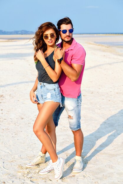 Outdoor fashion portrait of young pretty couple in love posing at amazing beach, wearing bright stylish casual clothes and sunglasses, enjoy their summer vacation near ocean.