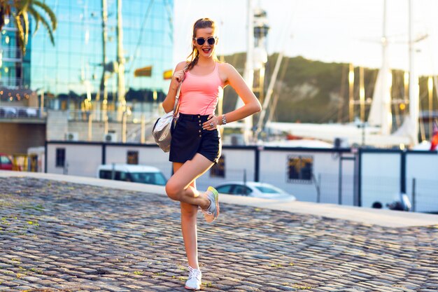 Outdoor fashion portrait of young sportive woman having fun travel and walking at luxury yacht club, evening sunlight, bright colors