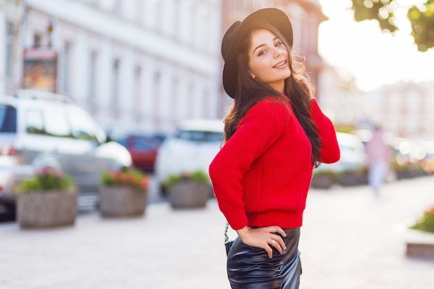 Внешнее изображение уличного стиля моды соблазнительной женщины брюнетки в осеннем случайном оборудовании, идущем в солнечном городе. Красный вязаный пуловер, черная модная шапка, кожаная юбка.