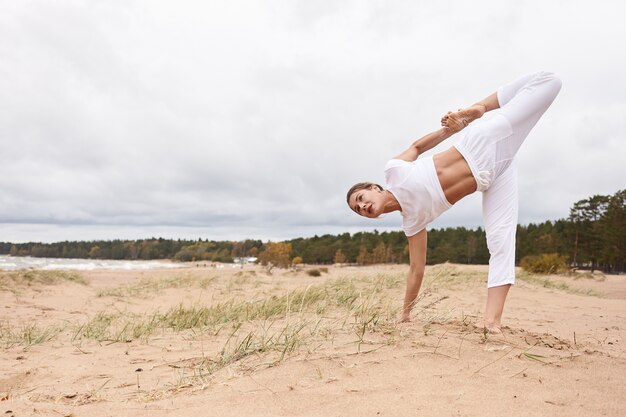 Immagine a figura intera all'aperto di una donna caucasica concentrata che indossa un abito bianco, facendo yoga all'aperto, in piedi con un piede e una mano sulla sabbia, allenamento dell'equilibrio, concentrazione e coordinazione