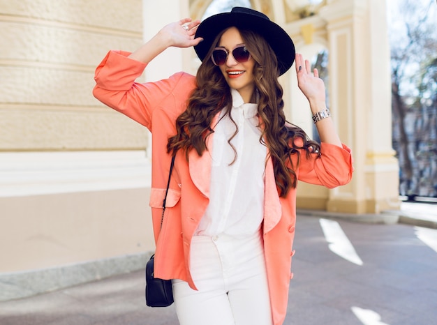 Outdoor hight fashion portrait of sexy stylish casual woman in black hat, pink suit, white blouse posing on old street. Spring, fall sunny day. Wavy hairstyle.