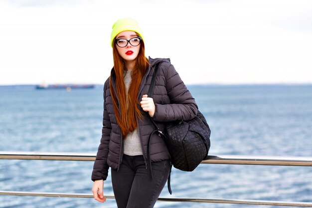 outdoor hipster fashion portrait of young pretty woman with long ginger hairs, posing near seaside, traveling alone with backpack,cold weather, stylish street style winter look, hat, jacket, sweater.