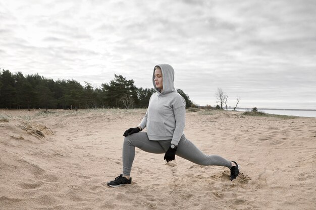 砂浜で運動し、突進し、走る前に体を温めるトレンディなスポーツウェアの魅力的なスリムで筋肉質の若い女性アスリートの屋外画像。スポーツ、フィットネス、柔軟性、強さ