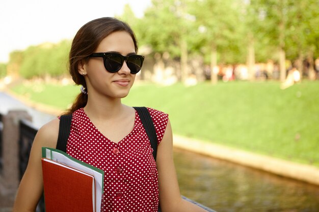 Outdoor image of cheerful female student wearing stylish black sunglasses and red dotted dress smiling, having cheerful look, carrying copybooks under her arm on her way to university in the morning