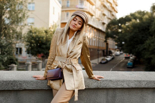 秋の街を歩くエレガントなヨーロッパの女性の屋外イメージ。ベージュのキャップとジャケット。スタイリッシュなアクセサリー。