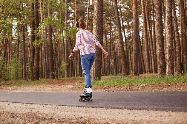 Outdoor image of energetic sleder young woman having training, being active, rollerblading alone, having rest, relaxing, enjoying weekends, being on road, listening to music. Lifestyle concept.