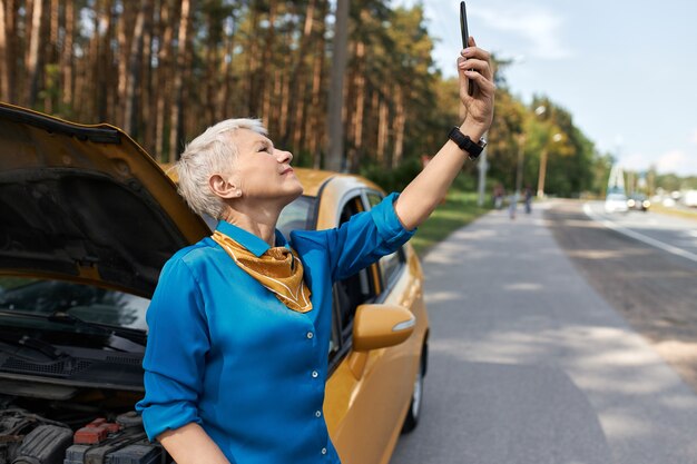 Outdoor image of unhappy middle aged woman standing on road by broken car with open hood raising hand with mobile phone, looking for network signal, trying to call for assistance.