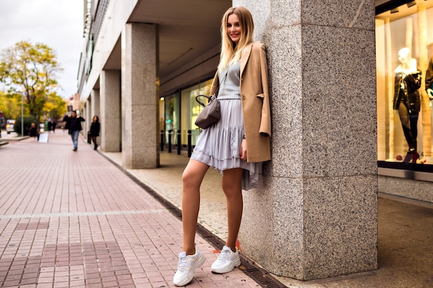 Outdoor lifestyle fashion portrait of pretty elegant glamour blonde woman with long legs, wearing trendy sneakers, dress sweater and coat, posing at Europe city, traveling alone.