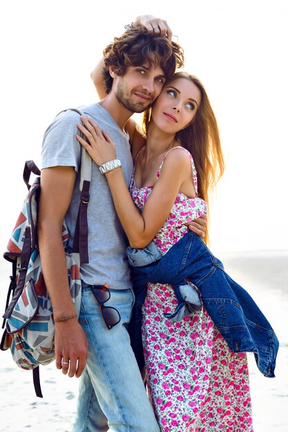 Outdoor lifestyle portrait of amazing pretty young couple in love posing on the beach. Stylish man and woman hugs and spend great time together. floral dress backpack and denim.