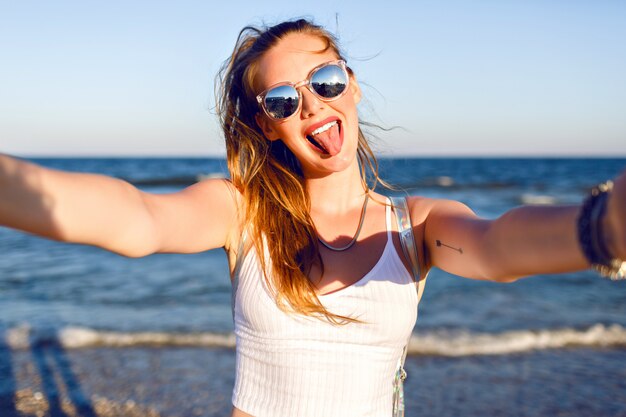 Outdoor lifestyle portrait of funny happy girl traveling to the ocean alone, making selfie at the beach, happy positive emotions, mirrored sunglasses, white crop top and backpack, joy, motion.