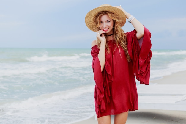 Outdoor lifestyle portrait of pretty white ginger woman in stylish dress posing on the beach near ocean. Blu sky. Windy weather.