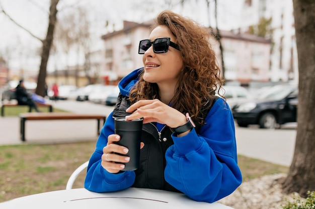 Outdoor photo of attractive curly woman in sunglasses wearing bright blue pullover and jacket is drinking coffee to go on summer terrace in warm spring day