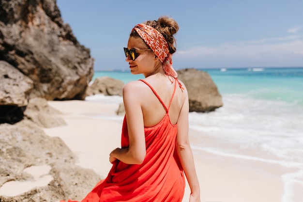 魅力的な日焼けした女の子の後ろからの屋外写真は、流行のリボンを着ています。海の近くの野生のビーチでリラックスした赤いドレスを着た壮大な若い女性の肖像画。