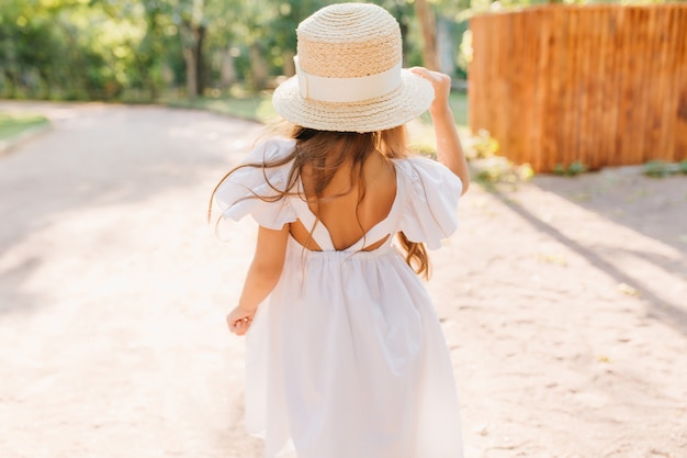 晴れた朝、通りに立っている日焼けした肌の少女の後ろからの屋外写真。魅力的な女性の子供は、公園で踊るリボンと白いドレスで飾られた麦わら帽子をかぶっています。