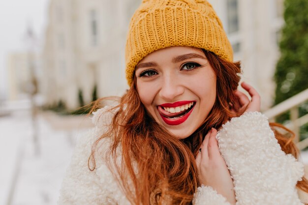 겨울을 즐기는 장난 생강 여자의 야외 사진. 야외 산책하는 노란 모자에 사랑스러운 아가씨를 웃 고있다.