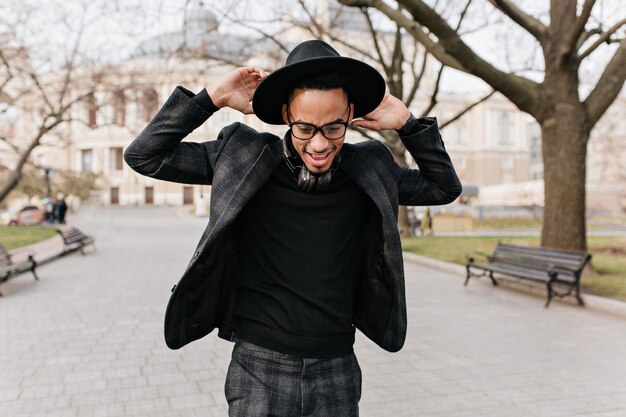 Outdoor photo of slim african man having fun in spring park. Portrait of enthusiastic brunette black guy in hat dancing on the street.