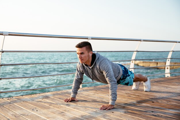 Outdoor photo of a sportive man in headphones doing push-ups exercises, during workout