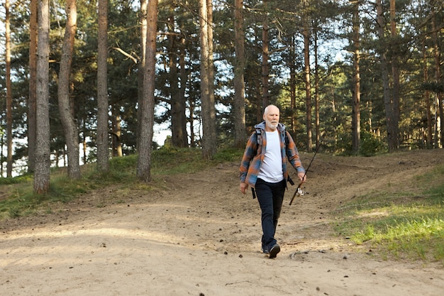 Outdoor picture of sad elderly bearded man with fishing rod going along path in woods, having disappointed facial expression because he caught no fish at all. on fishing spot Activity and recreation