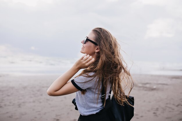 Outdoor portrait of amazing girl with long dark hair expressing happiness during walk around beach. Inspired female model in gray t-shirt spending time near sea in cloudy day.