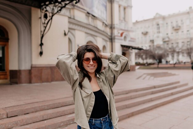 Outdoor portrait of attractive dark haired woman wearing stylish sunglasses and denim jacket walking on spring city