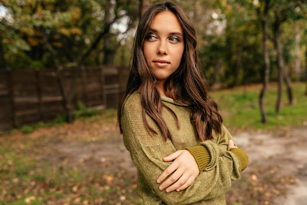 Free photo outdoor portrait of attractive young woman with long wavy hairstyle is wearing green pullover is looking aside with lovely smile with crossed hands on chest