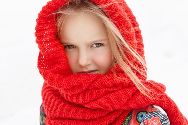 Outdoor portrait of beautiful blonde Caucasian little girl wrapped in warm red scarf