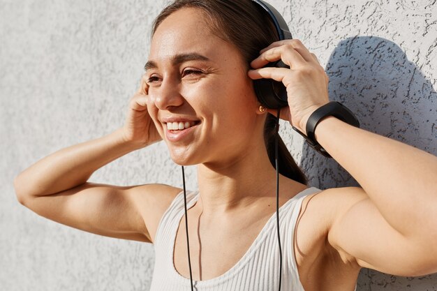 Outdoor portrait of beautiful satisfied positive happy female wearing white top, listening music