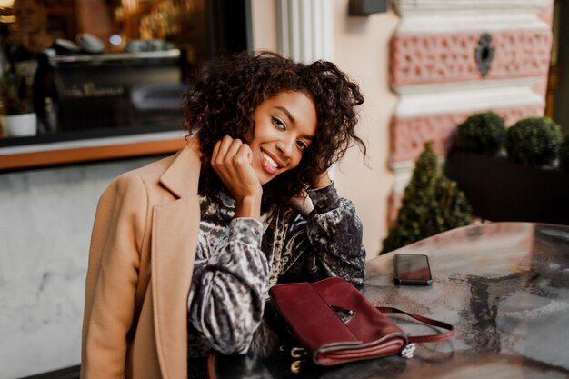 Outdoor  portrait of  beautiful  smiling black woman with stylish afro hairs sitting in cafe in Paris.