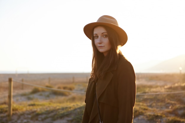 Outdoor portrait of beautifulCaucasian woman wearing fashionable coat and hat feeling carefree and peaceful, contemplating amazing morning view at seaside, looking with charming smile