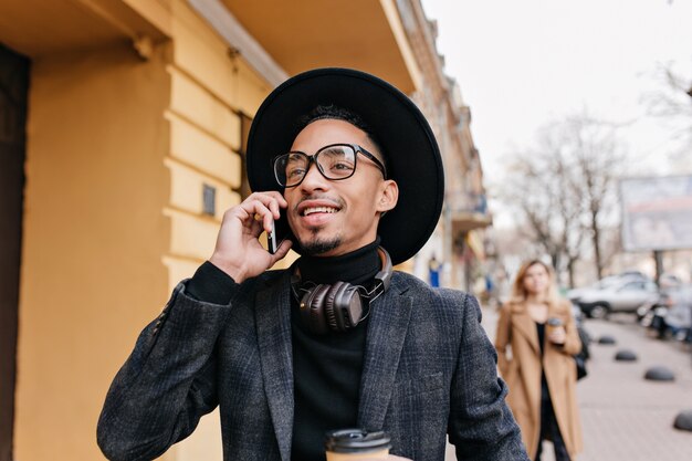 전화 통화 하 고 멀리보고 꿈꾸는 갈색 머리 남자의 야외 초상화. 도시 거리에 누군가 호출 바쁜 웃는 아프리카 소년의 사진.