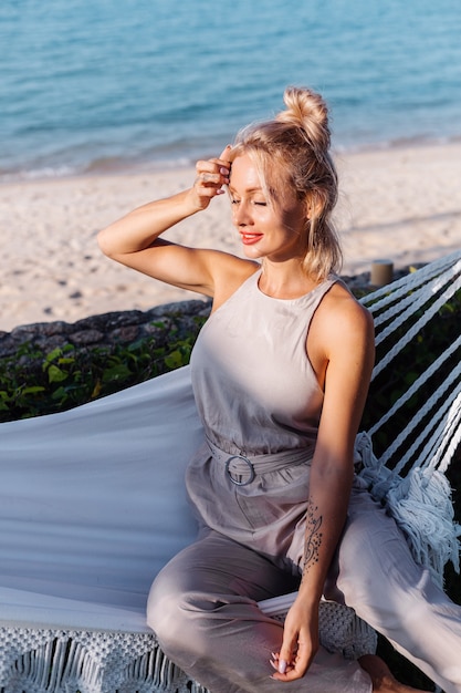 Outdoor portrait of caucasian woman in classic jumpsuit with red lipstick by hammock on vacation outside villa hotel, sea side