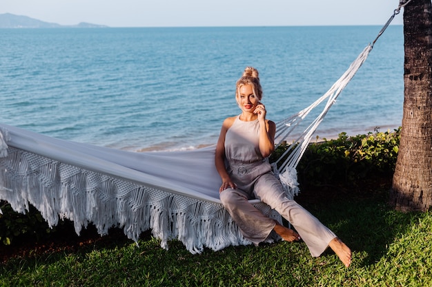 Outdoor portrait of caucasian woman in classic jumpsuit with red lipstick by hammock on vacation outside villa hotel, sea side