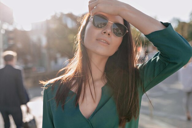 Открытый портрет очаровательной молодой женщины с каштановыми волосами в солнцезащитных очках касается ее волос и позирует перед камерой при солнечном свете в городе