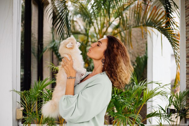 Outdoor portrait of curly european tanned woman holds happy pet dog pomeranian spitz