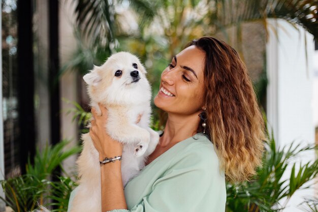 Outdoor portrait of curly european tanned woman holds happy pet dog pomeranian spitz