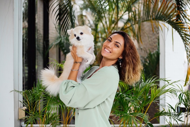 Outdoor portrait of curly european tanned woman holds happy pet dog pomeranian spitz