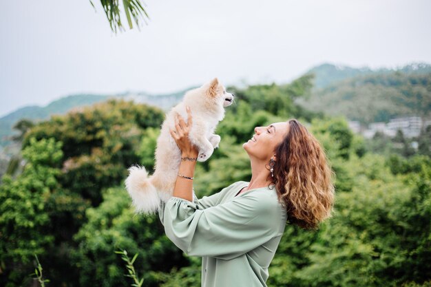 Outdoor portrait of curly european tanned woman holds happy pet dog pomeranian spitz