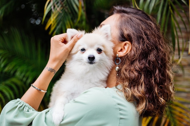巻き毛のヨーロッパの日焼けした女性の屋外の肖像画は幸せなペットの犬のポメラニアンスピッツを保持します