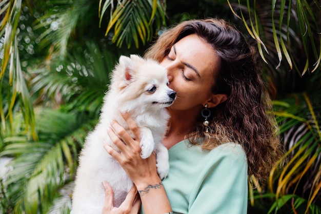 Outdoor portrait of curly european tanned woman holds happy pet dog pomeranian spitz