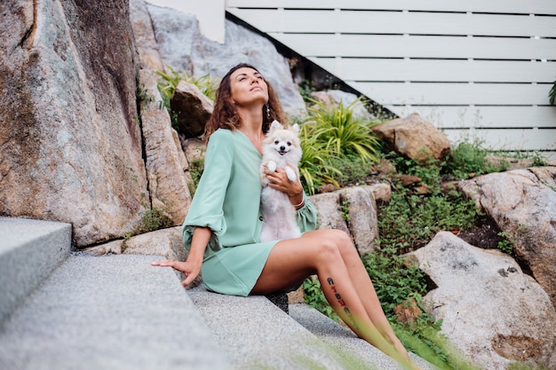 Outdoor portrait of curly european tanned woman holds happy pet dog pomeranian spitz