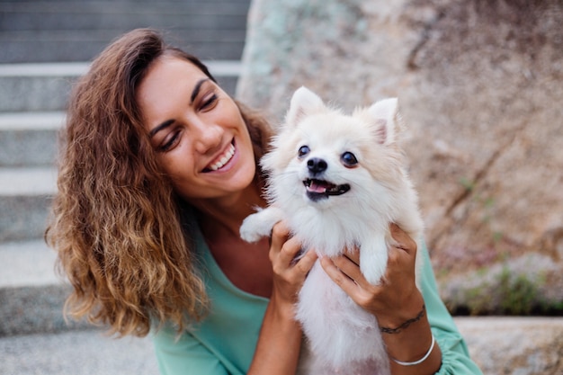 Outdoor portrait of curly european tanned woman holds happy pet dog pomeranian spitz