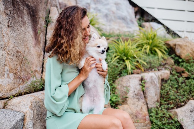 Outdoor portrait of curly european tanned woman holds happy pet dog pomeranian spitz