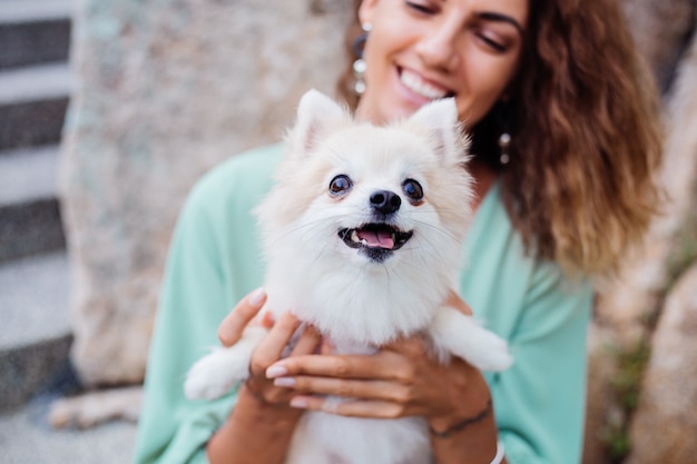 Outdoor portrait of curly european tanned woman holds happy pet dog pomeranian spitz