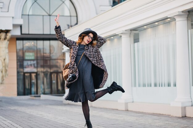 Outdoor portrait of elegant young lady with brown backpack wearing coat and hat. Attractive woman with curly hair speaking jumping and having fun.