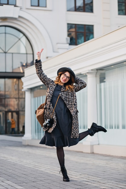 Outdoor portrait of elegant young lady with brown backpack wearing coat and hat. Attractive woman with curly hair speaking jumping and having fun.