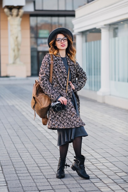 Outdoor portrait of elegant young lady with brown backpack wearing coat and hat. Attractive woman with curly hair speaking on phone while drinking coffee on the street and waiting friends..